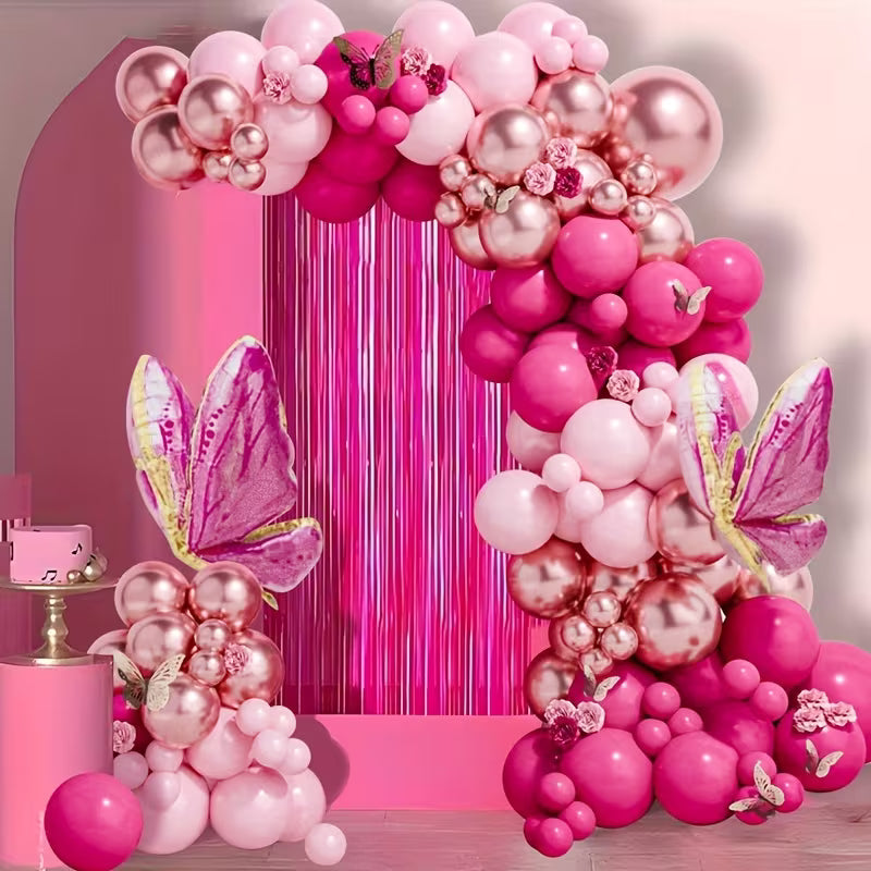 A pink and rose gold balloon garland decoration set featuring a large arch, a matching pedestal cluster, and two large butterfly balloons with glittery pink wings, set against a pink metallic fringe backdrop.